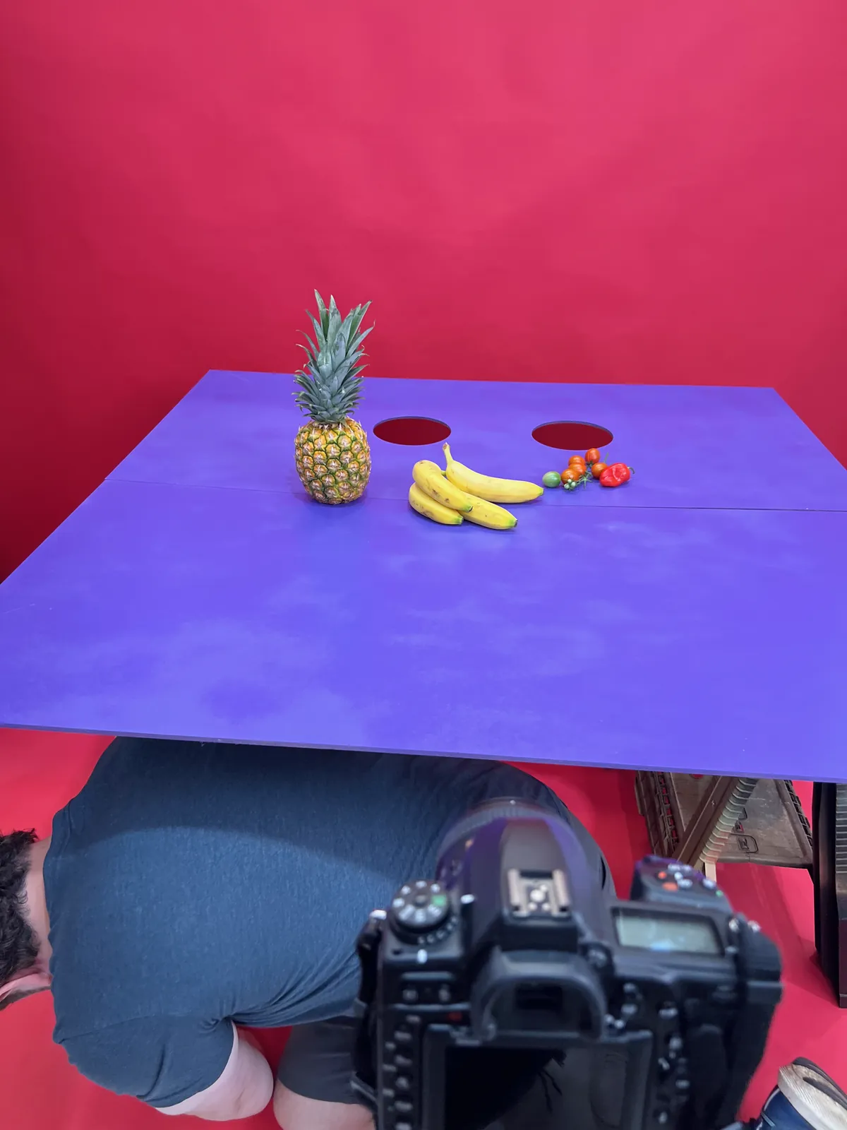 Close on the violet tabletop and its two hand holes, set with a pineapple and bananas for a test shot, with someone crouched underneath.