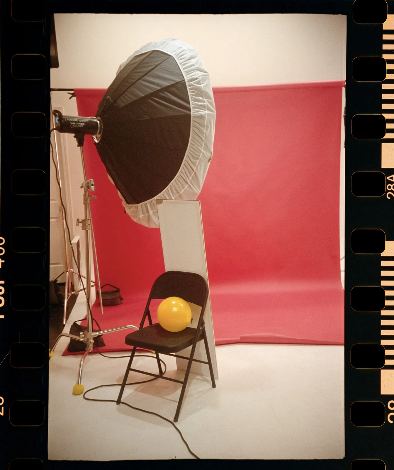 A film-frame snapshot of the studio: a folding chair holding a yellow balloon beside a large softbox.