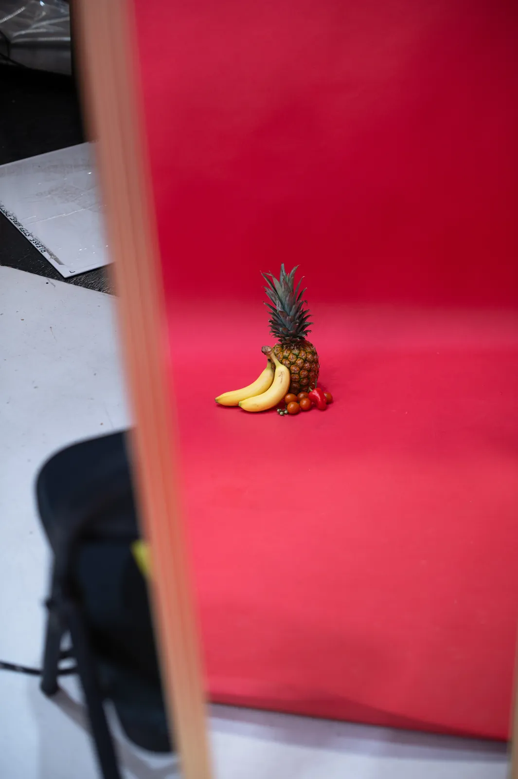 A pineapple, bananas and tomatoes posed alone on the red backdrop, seen past a light stand.