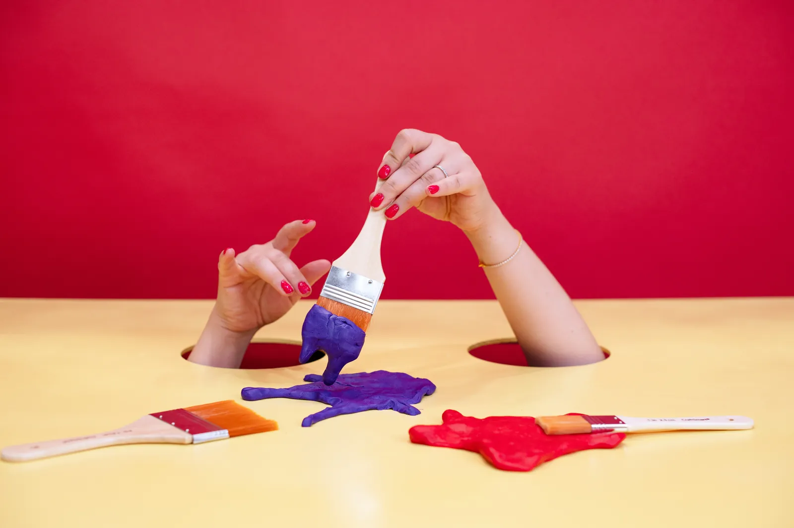 Two hands reach up through holes in a yellow tabletop, one lifting a paintbrush dripping violet clay paint over red and violet splats.