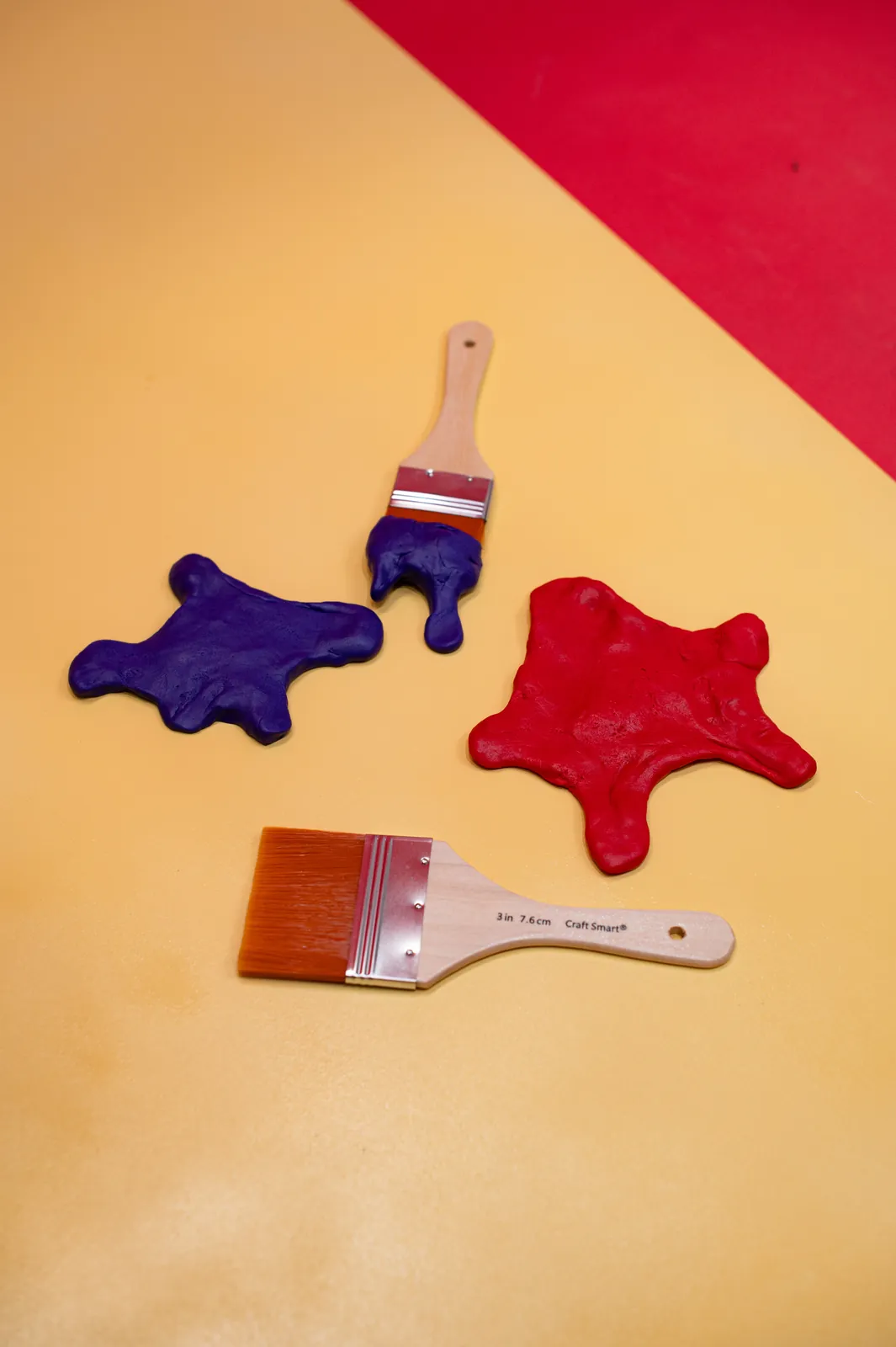 Overhead: red and violet clay paint splats and two brushes arranged on a yellow board against red.