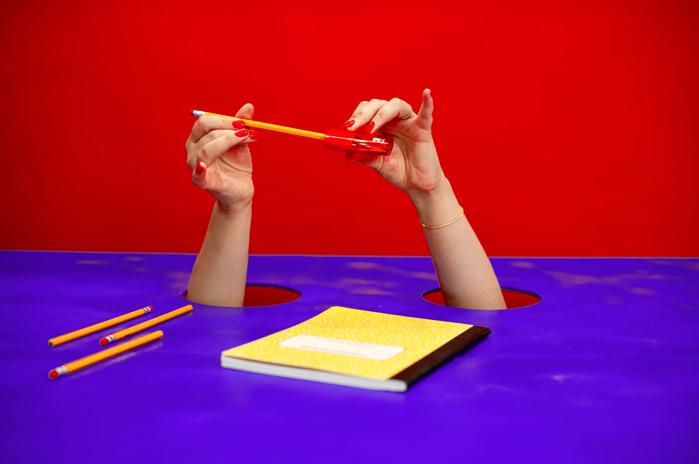 Hands hold a pencil up over a yellow notebook, with loose pencils on the violet table.