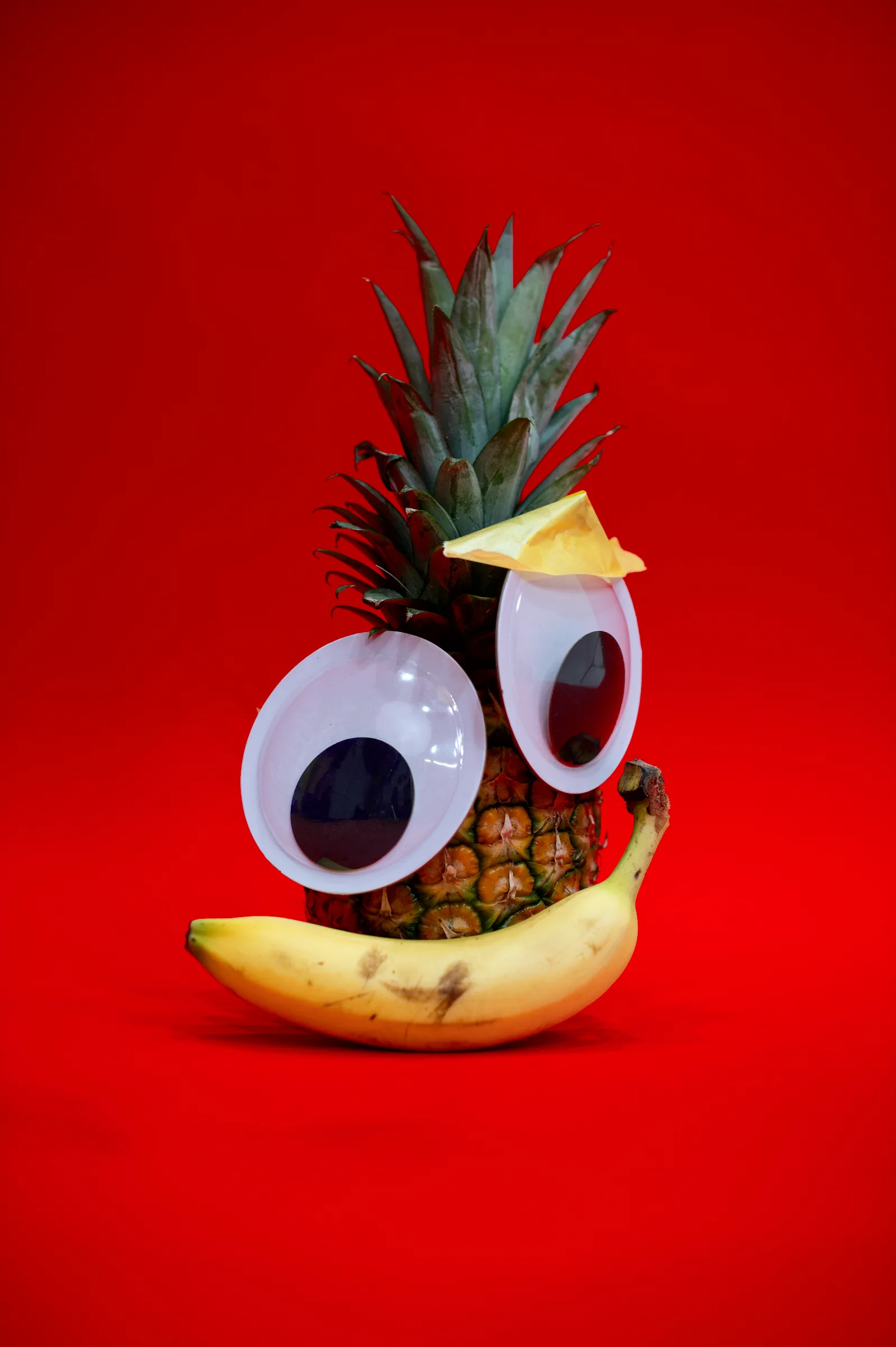 A pineapple with googly eyes and a banana smile, on red.