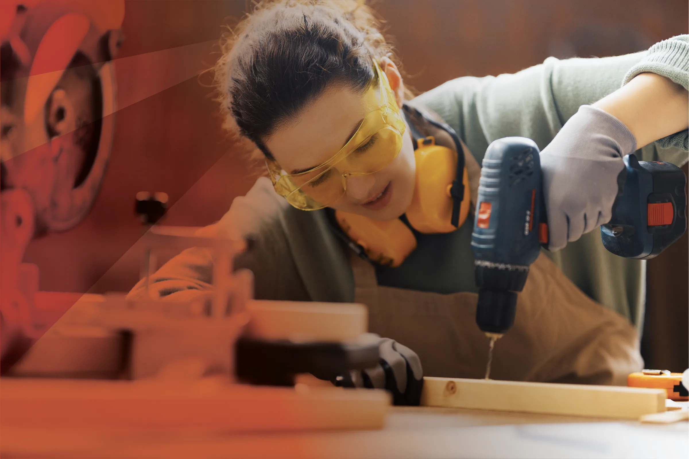 A young woman in safety goggles and ear defenders drills into a board in a workshop.